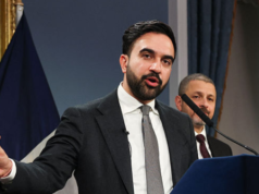 Mamdani tossisce in mano – In piedi accanto all’attivista che indossa la maschera